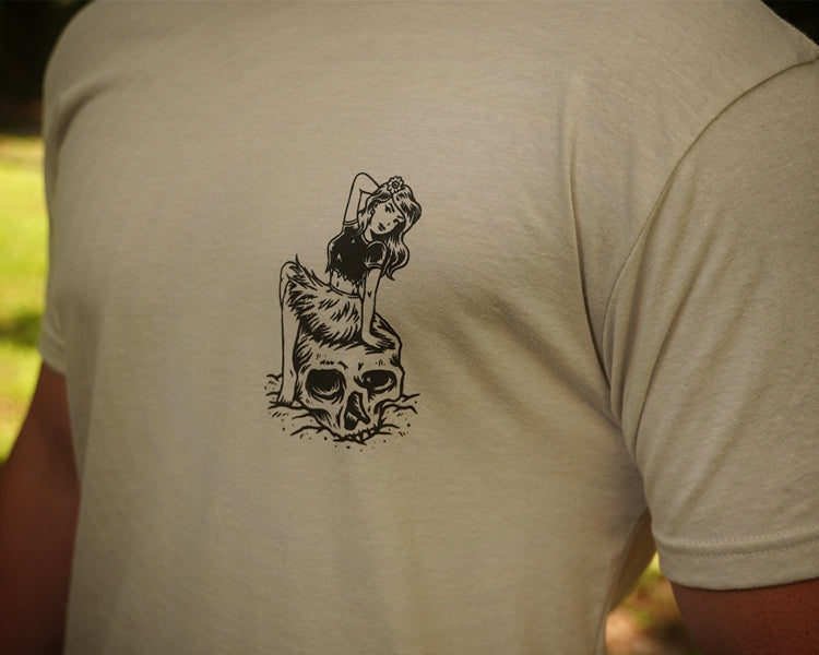 Person wearing a beige t-shirt with a black graphic design on the back, set against a blurred natural background.
