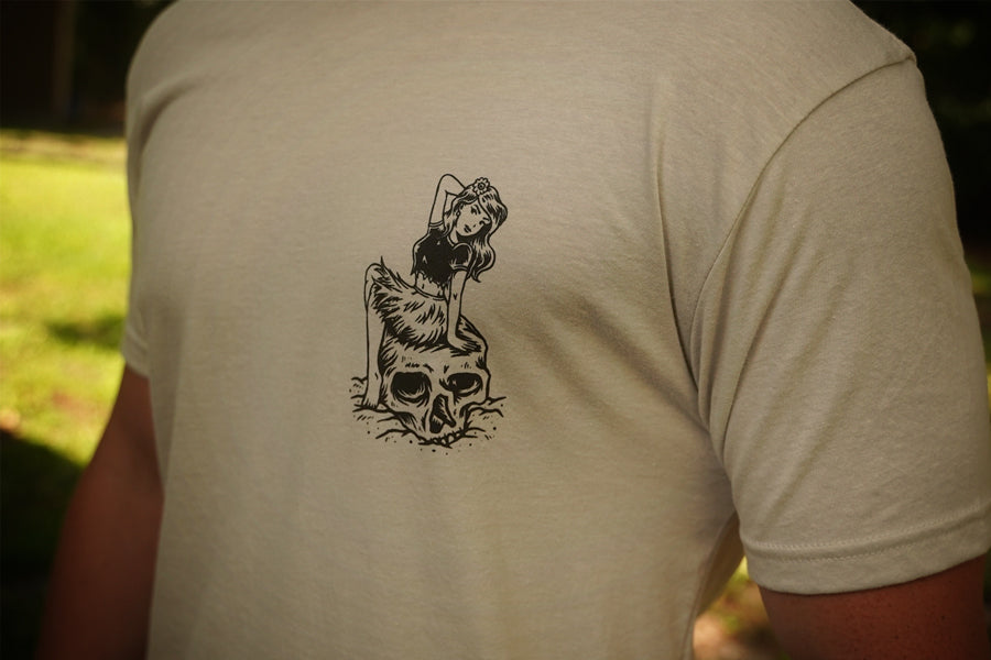 Person wearing a beige t-shirt with a black graphic design on the back, set against a blurred natural background.