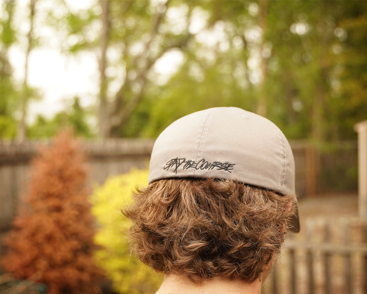 Person wearing a cap with Stay the Course, standing in a backyard with trees and a fence.