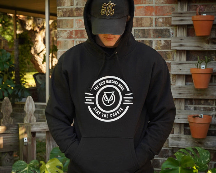 Person wearing a black hoodie with a logo and text, standing in front of a brick wall with potted plants.