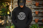 Person wearing a black hoodie with a logo and text, standing in front of a brick wall with potted plants.