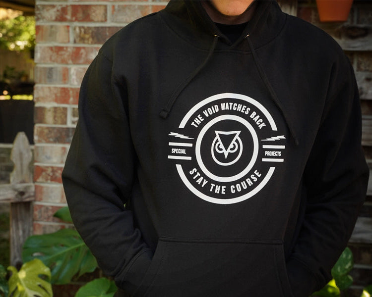 Person wearing a black hoodie with a white logo and text, standing in front of a brick wall.