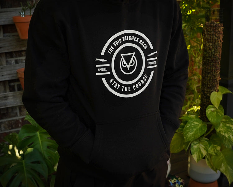 Black hoodie with a white logo and text worn by a person, set against a natural background with plants.