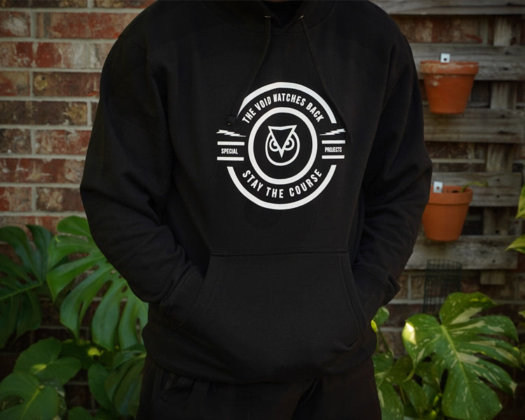 Person wearing a black hoodie with a white logo against a brick wall and plant background