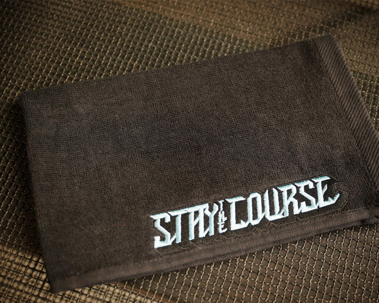 A black towel featuring the embroidered baby blue text 'STAY COURSE' in a stylized font, laid flat on a brown woven surface.