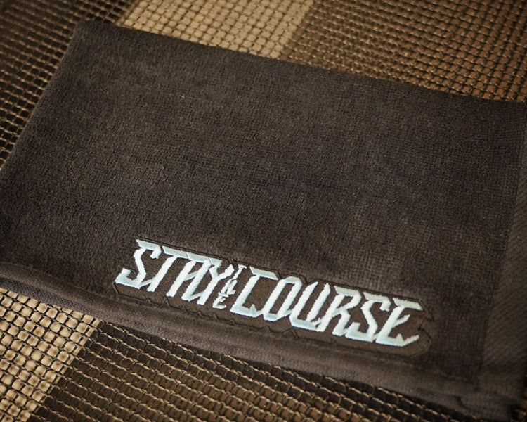 A black towel with the words 'STAY ON COURSE' embroidered in a stylized light blue font, resting on a textured mat.