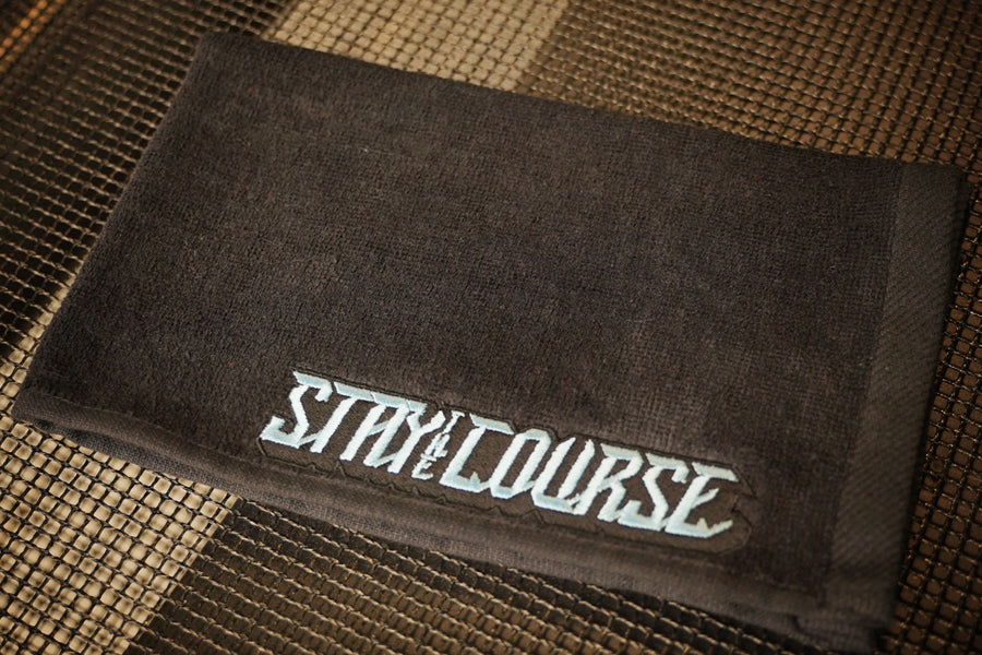 A black towel with the words 'STAY ON COURSE' embroidered in a stylized light blue font, resting on a textured mat.
