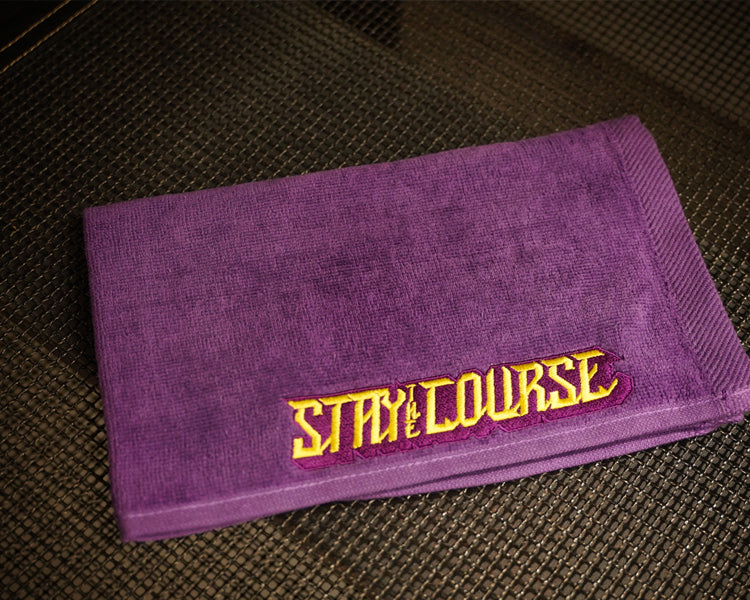 A purple towel with 'STAY ON COURSE' embroidered in yellow and purple resting on a dark textured surface.