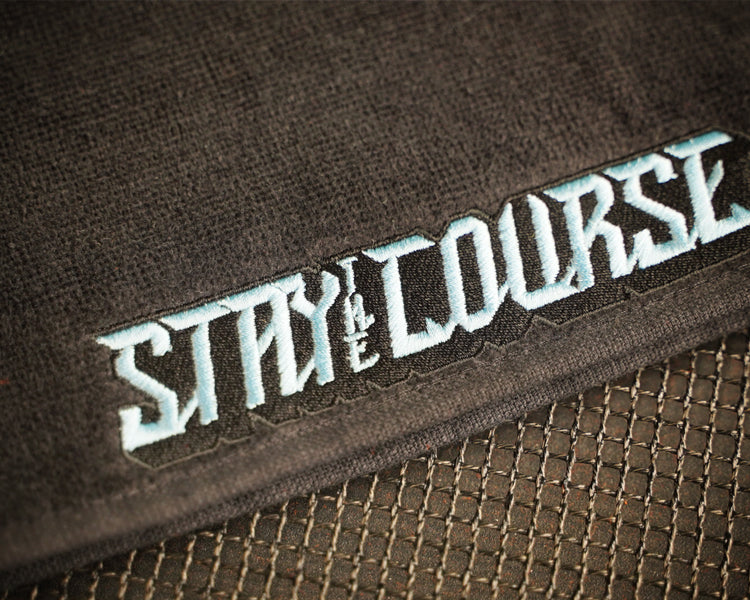 Close-up of embroidery with the words 'STAY ON THE COURSE' in stylized light blue lettering, placed over a dark mesh surface.