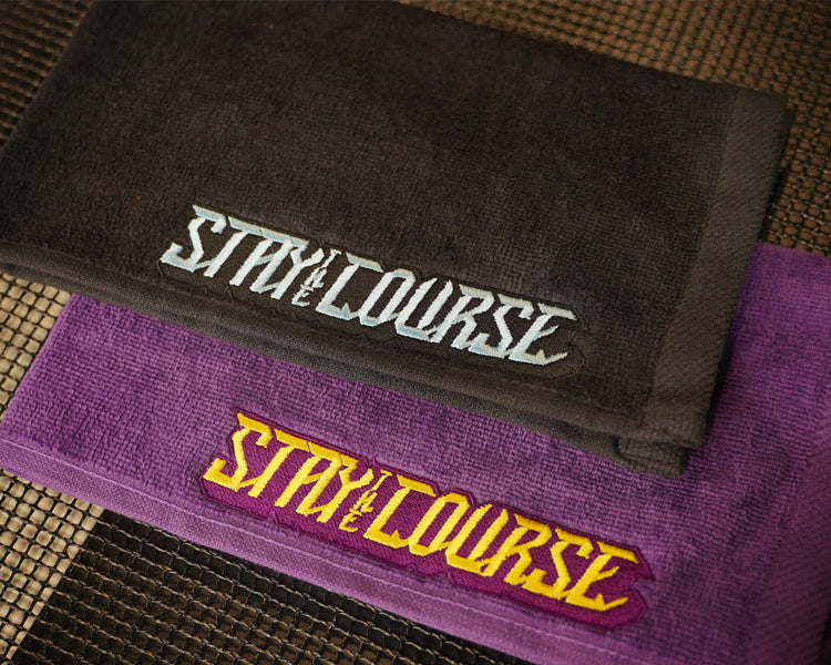 Two folded towels stacked on a woven mat: a black towel on top with baby blue embroidered 'STAY ON THE COURSE' text, and a purple towel underneath with yellow and purple embroidered 'STAY ON THE COURSE' text.