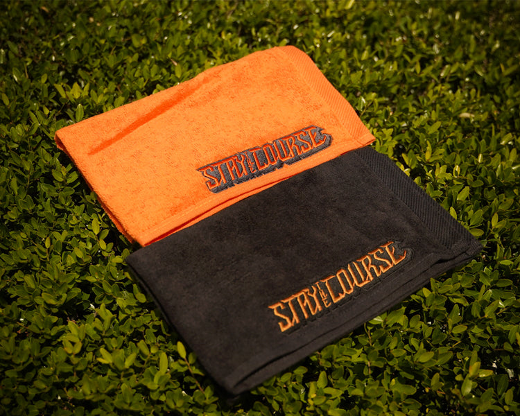 Two folded towels, one orange and one black, with 'Stay the Course' branding on a grassy background.