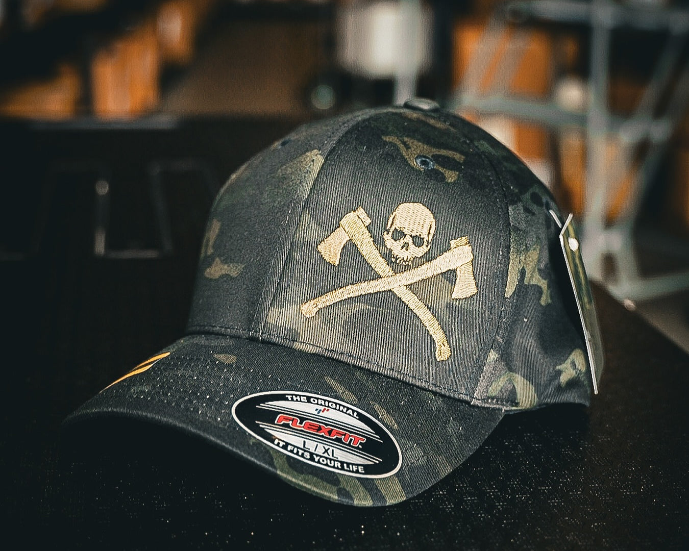 A dark camouflage baseball cap featuring a skull and crossed axes graphic on the front, resting on a dark surface with a blurred indoor background.