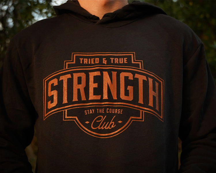 A close-up of a person wearing a black hoodie with a large rust colored graphic on the chest that reads 'TRIED & TRUE STRENGTH CLUB STAY THE COURSE'.