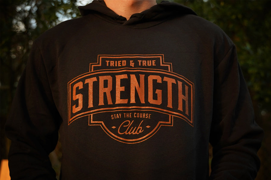 A close-up of a person wearing a black hoodie with a large rust colored graphic on the chest that reads 'TRIED & TRUE STRENGTH CLUB STAY THE COURSE'.