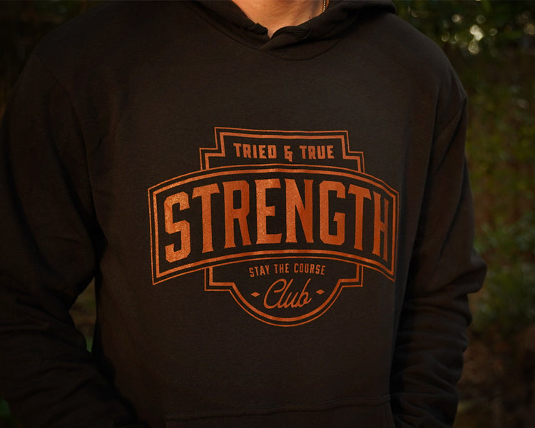 A person wearing a black hoodie with a large rust colored graphic on the chest that reads 'TRIED & TRUE STRENGTH CLUB' and 'STAY THE COURSE'. The background is a blurred outdoor scene with green foliage.
