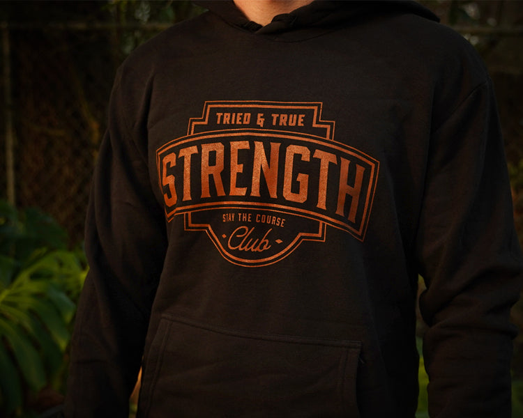 A person wearing a black hoodie with a large rust colored graphic on the chest that reads 'TRIED & TRUE STRENGTH CLUB' and 'STAY THE COURSE'. The background is a blurred outdoor scene with green foliage.