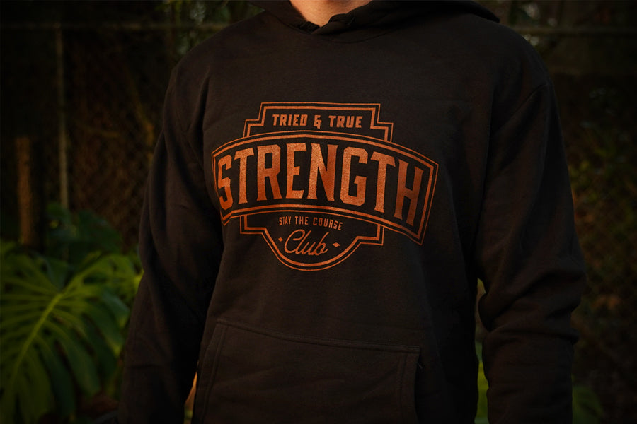 A person wearing a black hoodie with a large rust colored graphic on the chest that reads 'TRIED & TRUE STRENGTH CLUB' and 'STAY THE COURSE'. The background is a blurred outdoor scene with green foliage.