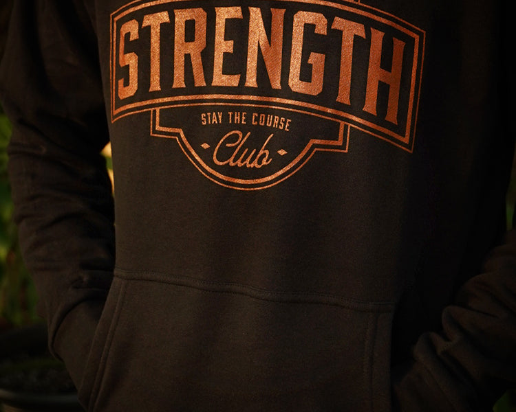 A close-up of a person wearing a black hoodie with a rust colored graphic print on the chest that reads 'STRENGTH Club' and 'STAY THE COURSE'.