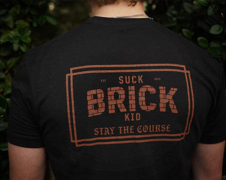 Person wearing a black t-shirt with 'Suck Brick Kid Stay the Course' text on the back, standing against a green leafy background.
