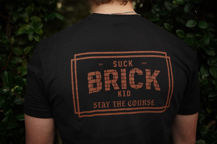 Person wearing a black t-shirt with 'Suck Brick Kid Stay the Course' text on the back, standing against a green leafy background.