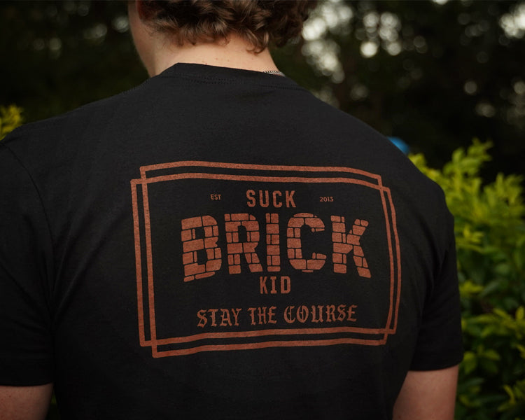Person wearing a black t-shirt with 'SUCK BRICK KID' text on the back, outdoors.