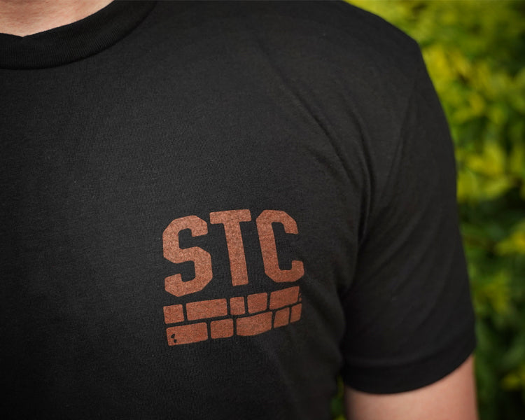 Black t-shirt with 'STC' logo worn by a person, against a blurred green background