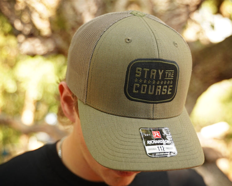 Olive cap with 'Stay the Course' embroidery worn by a person outdoors