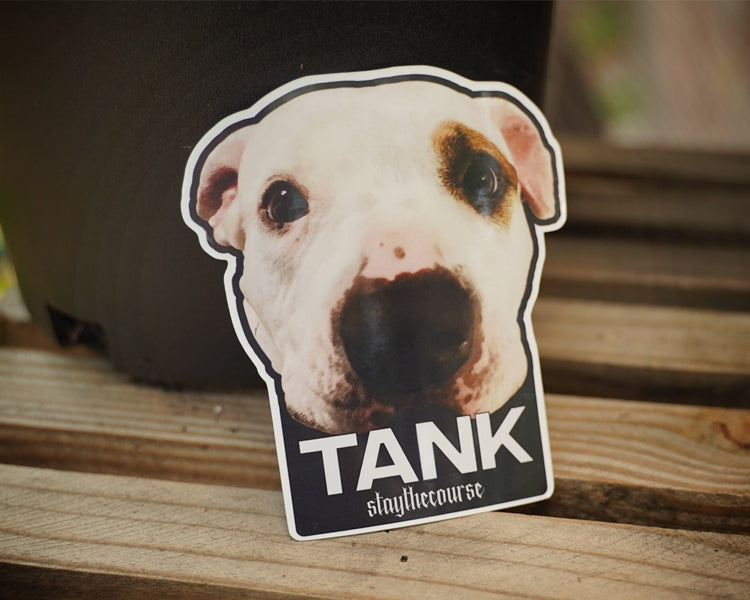Sticker of a dog's face with 'TANK' and 'staythecourse' text on a wooden surface.