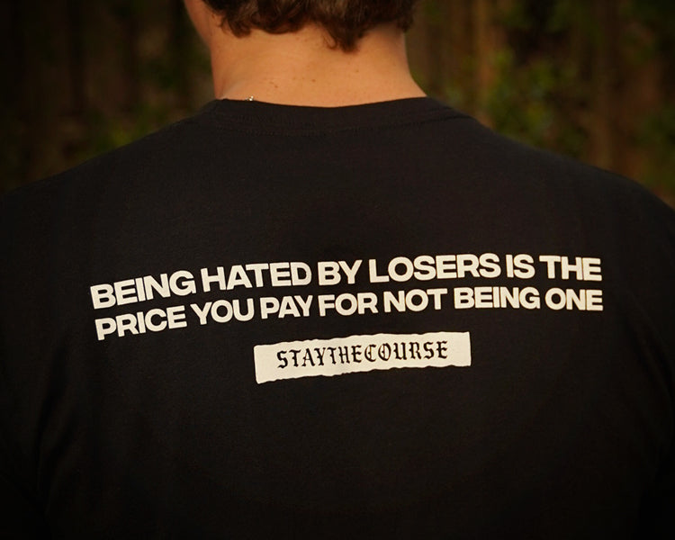 Person wearing a black t-shirt with white print that says "Being hated by losers is the price you pay for not being one, Stay the Course", in an outdoor setting