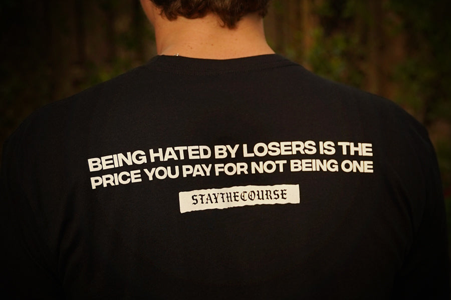Person wearing a black t-shirt with white print that says "Being hated by losers is the price you pay for not being one, Stay the Course", in an outdoor setting