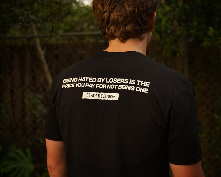 Person wearing a black t-shirt with white print that says "Being hated by losers is the price you pay for not being one, Stay the Course", in an outdoor setting
