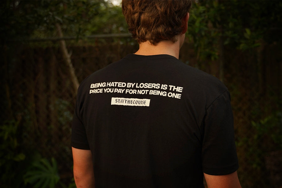 Person wearing a black t-shirt with white print that says "Being hated by losers is the price you pay for not being one, Stay the Course", in an outdoor setting