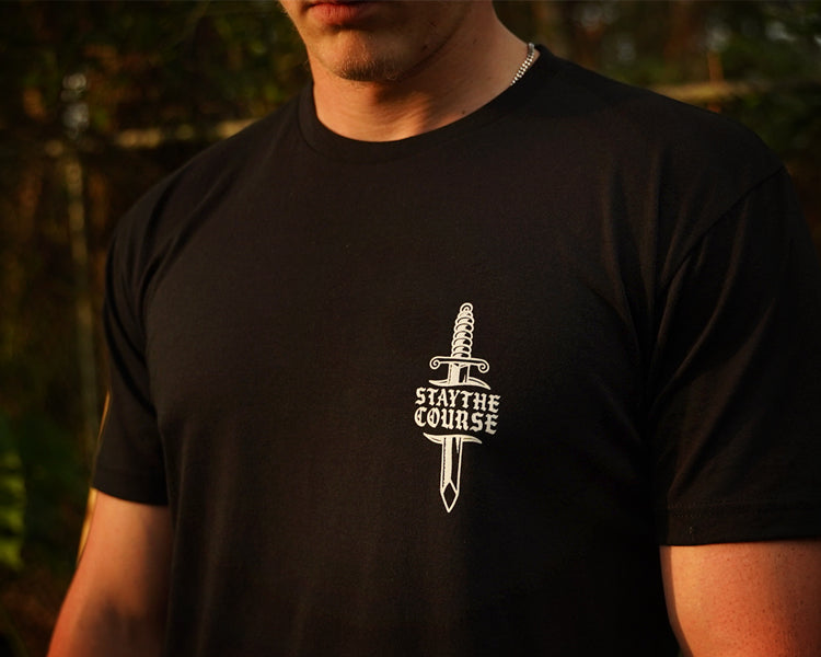 A person wearing a black t-shirt with what print of dagger and Stay the Course on the chest, in an outdoor setting