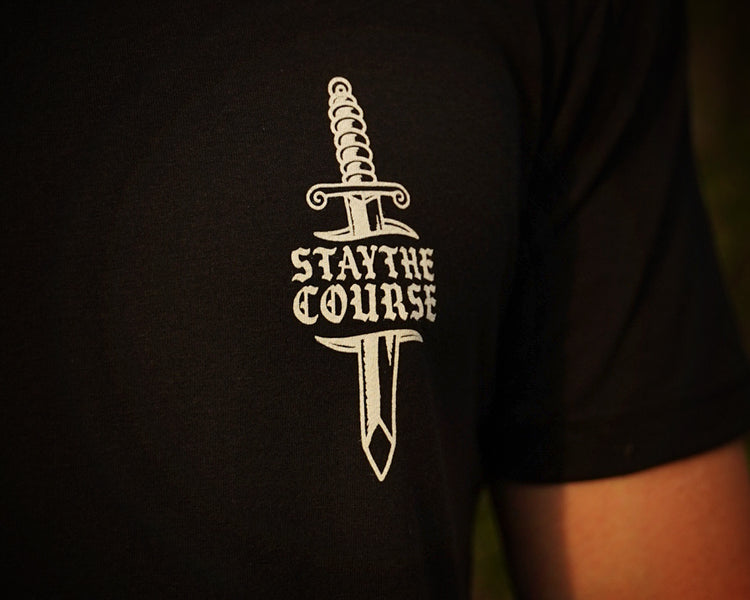 A close-up of a person wearing a black t-shirt with a white graphic of a dagger and the text 'STAY THE COURSE' printed on the chest.