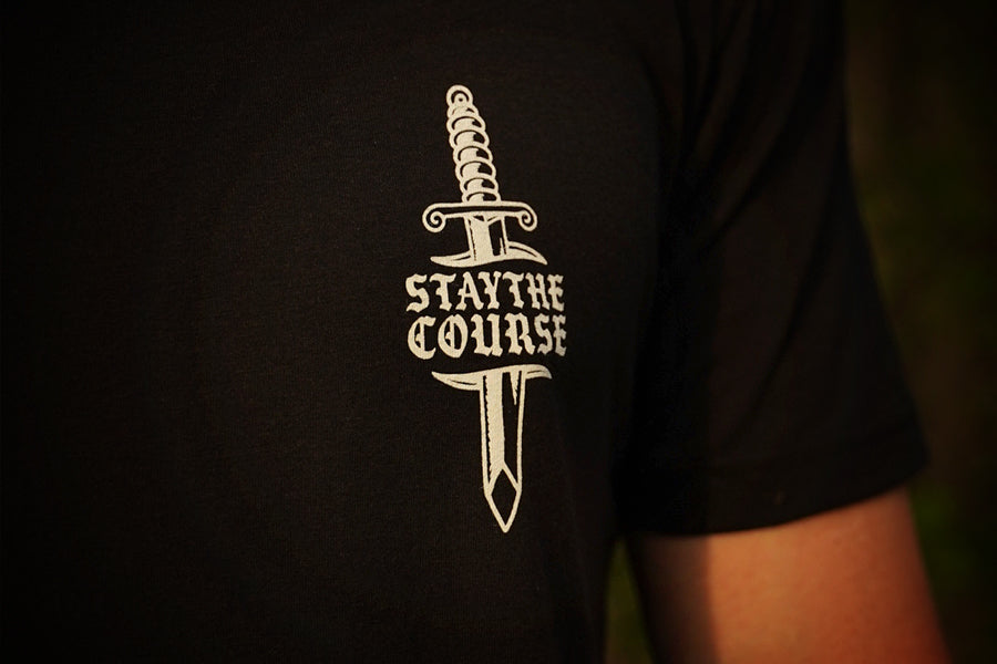 A close-up of a person wearing a black t-shirt with a white graphic of a dagger and the text 'STAY THE COURSE' printed on the chest.