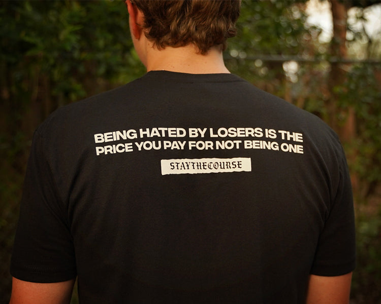 Person wearing a black t-shirt with white print that says "Being hated by losers is the price you pay for not being one, Stay the Course", in an outdoor setting