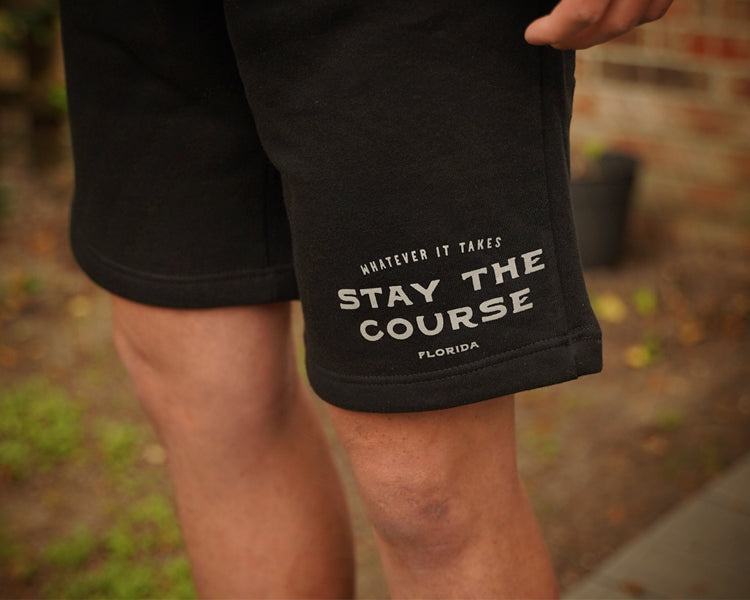 Black shorts with text 'Stay the Course Florida' held by a person outdoors.