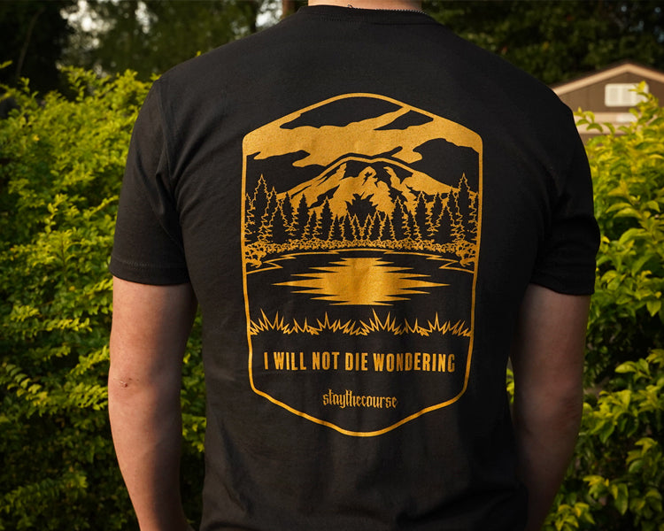 Person wearing a black t-shirt with a gold graphic and text in an outdoor setting