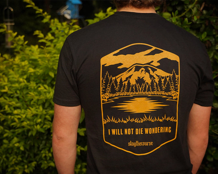 Person wearing a black t-shirt with a mountain and lake design on the back, standing in front of green foliage.