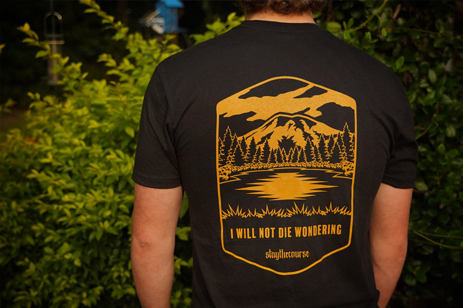 Person wearing a black t-shirt with a mountain and lake design on the back, standing in front of green foliage.
