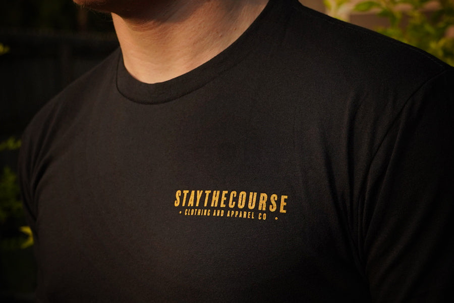 Black t-shirt with 'STAYTHECOURSE' logo on a blurred background