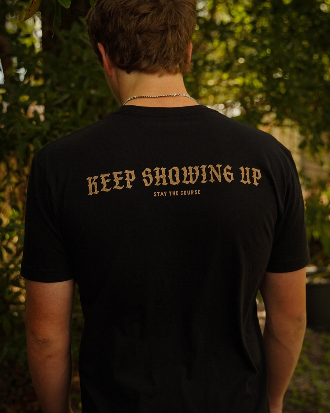 A person wearing a black T-shirt with white print on the back of a snake wrapped around a spear, with the words “ACTION OPENS DOORS” and “STAY THE COURSE”, set against a blurred outdoor background 