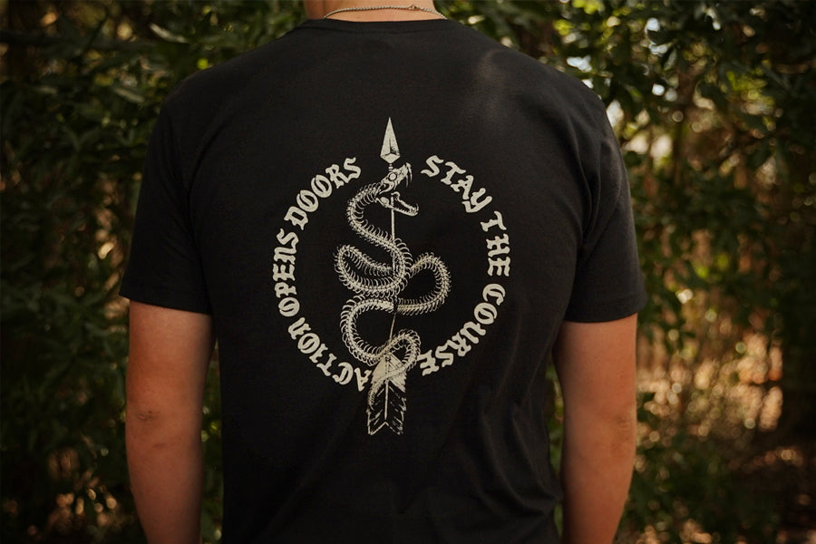 A person viewed from behind wearing a black short-sleeve T-shirt with a large white graphic print on the back. The design shows a coiled snake wrapped around a vertical spear, with the words “ACTION OPENS DOORS” on the left and “STAY THE COURSE” on the right, set against a blurred outdoor background of green foliage.