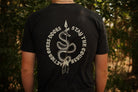 A person viewed from behind wearing a black short-sleeve T-shirt with a large white graphic print on the back. The design shows a coiled snake wrapped around a vertical spear, with the words “ACTION OPENS DOORS” on the left and “STAY THE COURSE” on the right, set against a blurred outdoor background of green foliage.