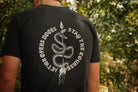 A person viewed from behind wearing a black T-shirt with a large white snake graphic and the text 'ACTION OPEN DOORS STAY THE COURSE' on the back, standing outdoors in front of blurred green foliage.