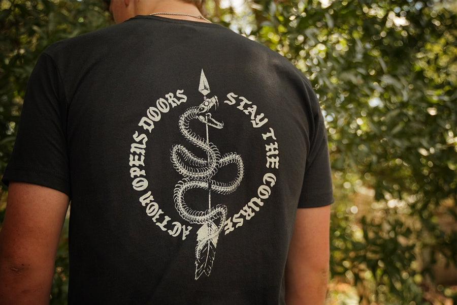 A person viewed from behind wearing a black T-shirt with a large white snake graphic and the text 'ACTION OPEN DOORS STAY THE COURSE' on the back, standing outdoors in front of blurred green foliage.