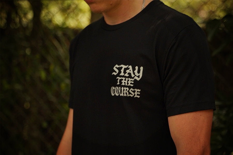 A person wearing a black t-shirt with white graphic that says STAY THE COURSE, standing against a blurred background