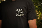A person wearing a black t-shirt with white graphic that says STAY THE COURSE, standing against a blurred background