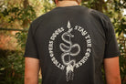 A person viewed from behind wearing a black T-shirt with a large white snake graphic and the text 'ACTION OPEN DOORS STAY THE COURSE' on the back, standing outdoors in front of blurred green foliage.