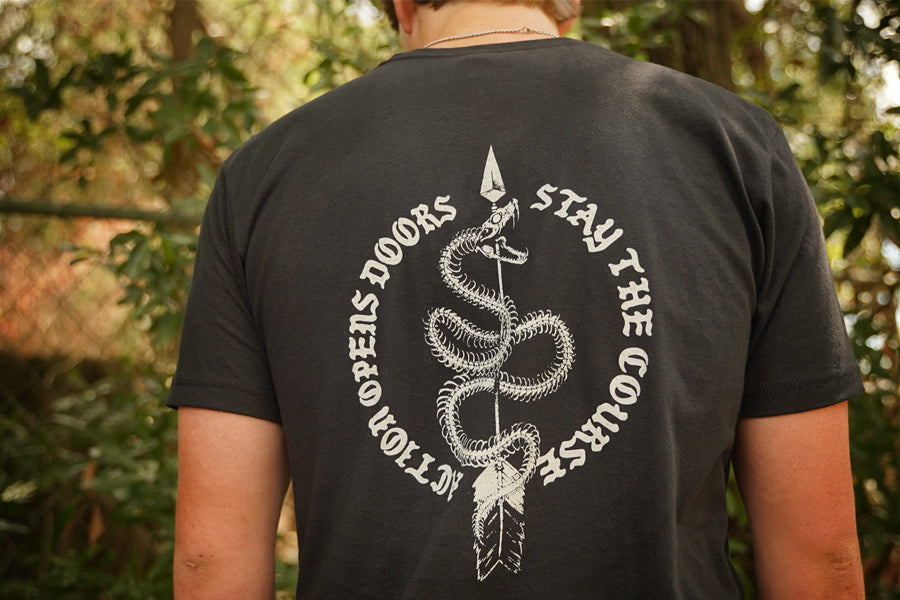 A person viewed from behind wearing a black T-shirt with a large white snake graphic and the text 'ACTION OPEN DOORS STAY THE COURSE' on the back, standing outdoors in front of blurred green foliage.
